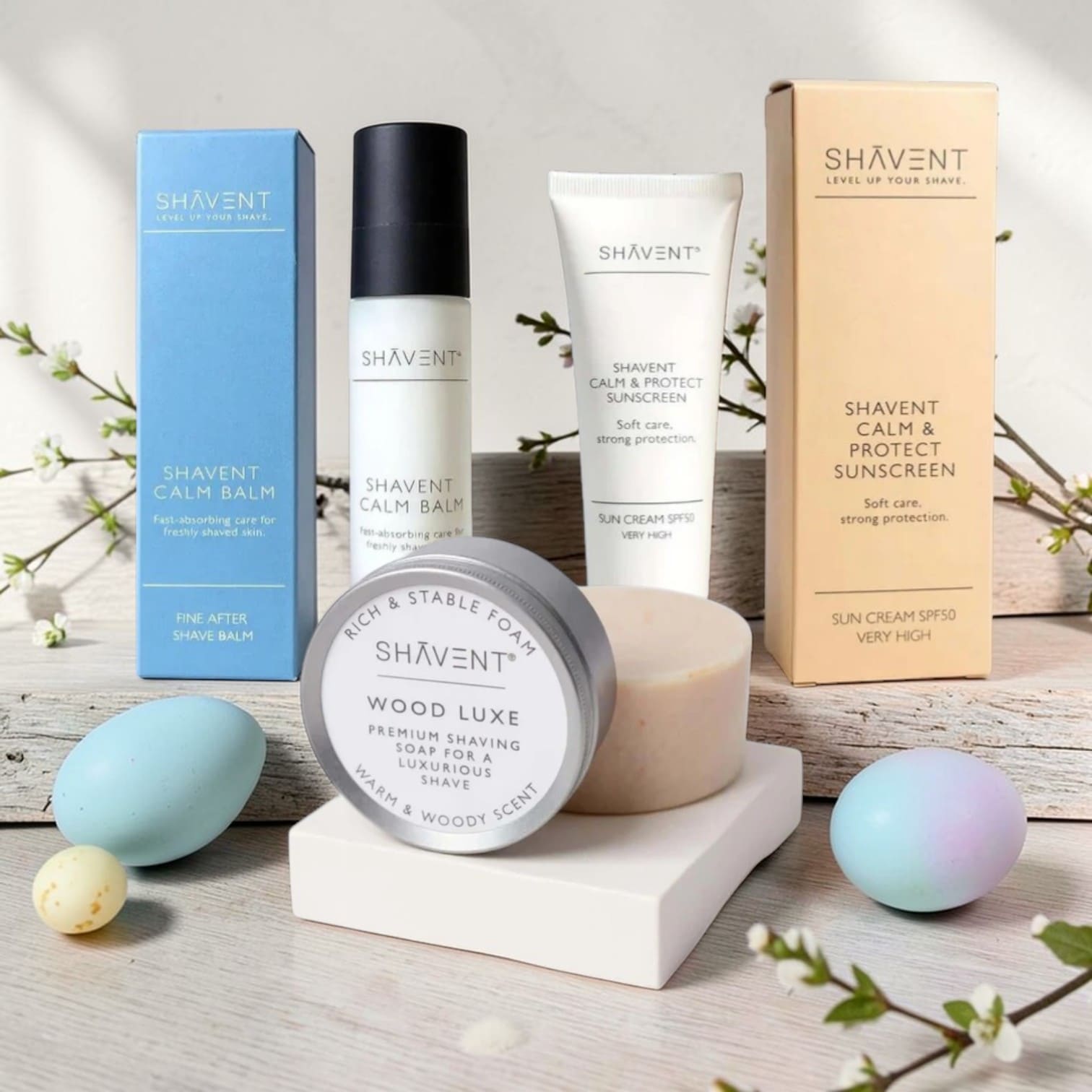 SHAVENT Grooming Oster-Special: Rasurpflege-Set mit Wood Luxe Rasierseife, Calm Balm und Calm & Protect Sonnencreme, arrangiert auf hellem Holz mit Frühlingsdeko.