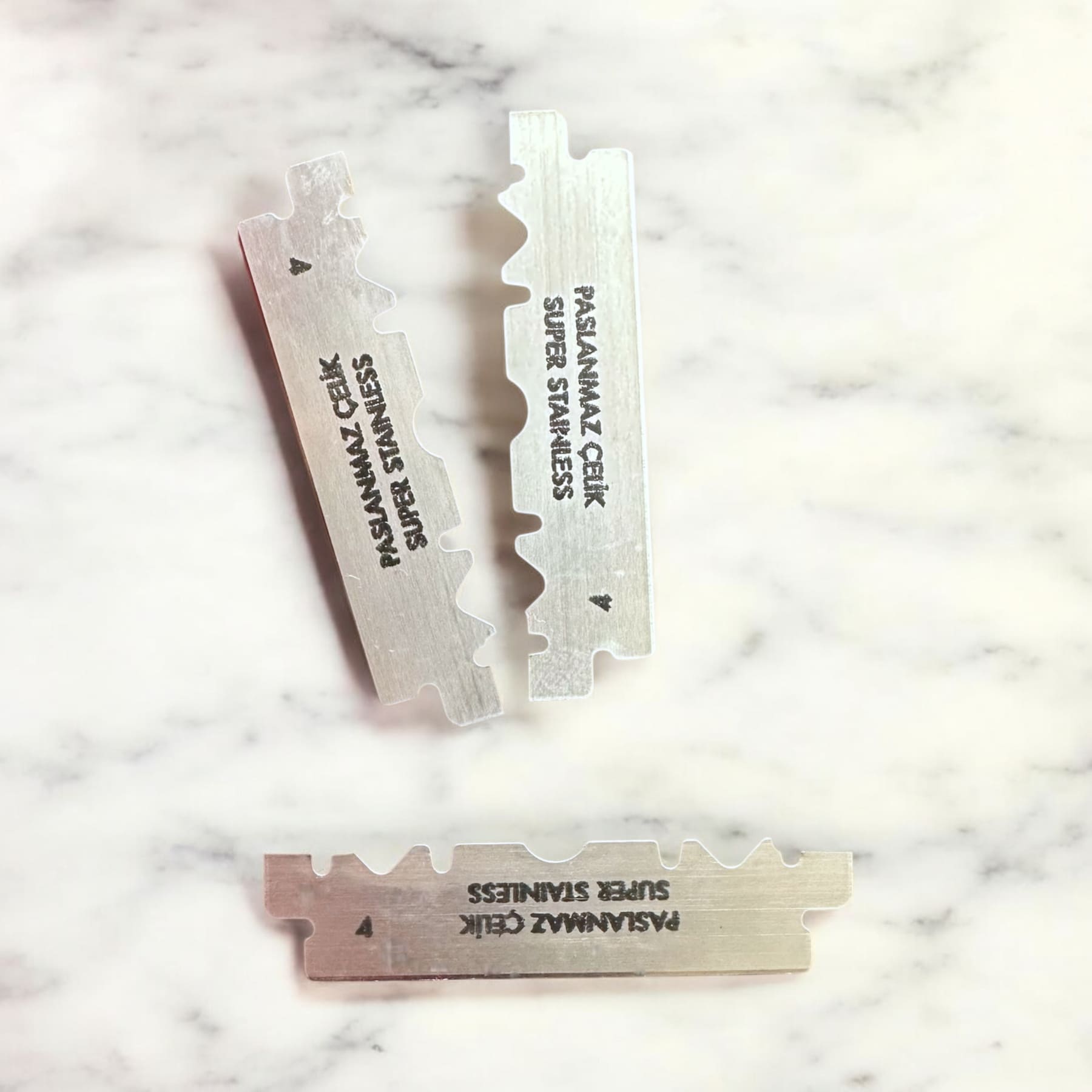 Drei doppelseitige PASANAMAZ ÇELİK Super Stainless Rasierklingen auf Marmor, Teil des SHAVENT Compact Oster Specials mit kostenlosem Klingen-Test-Set für sanfte Körperrasur.