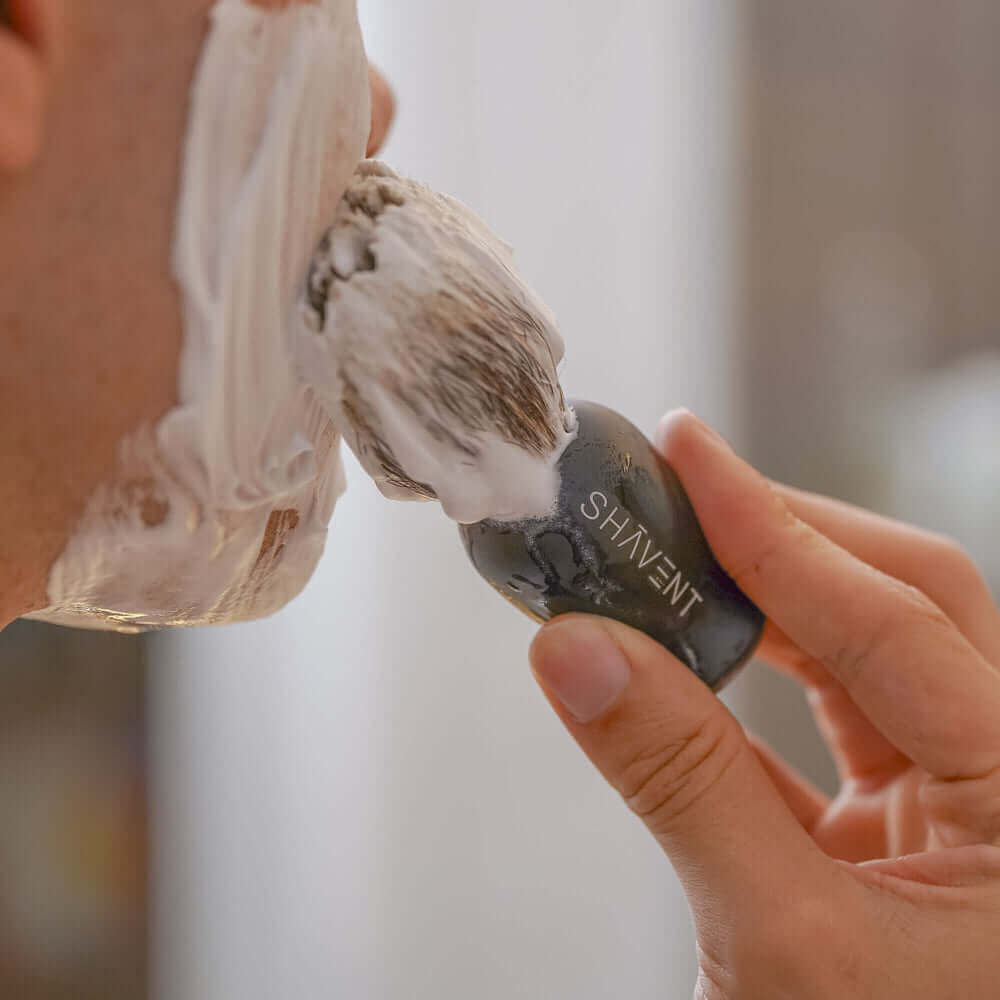 Person schäumt Gesicht mit SHAVENT Wood Luxe Rasierseife ein, hält schwarzen Rasierhobel. Hochwertige, pflegende Seife mit holzigem Duft, ideal für tägliche Rasur.