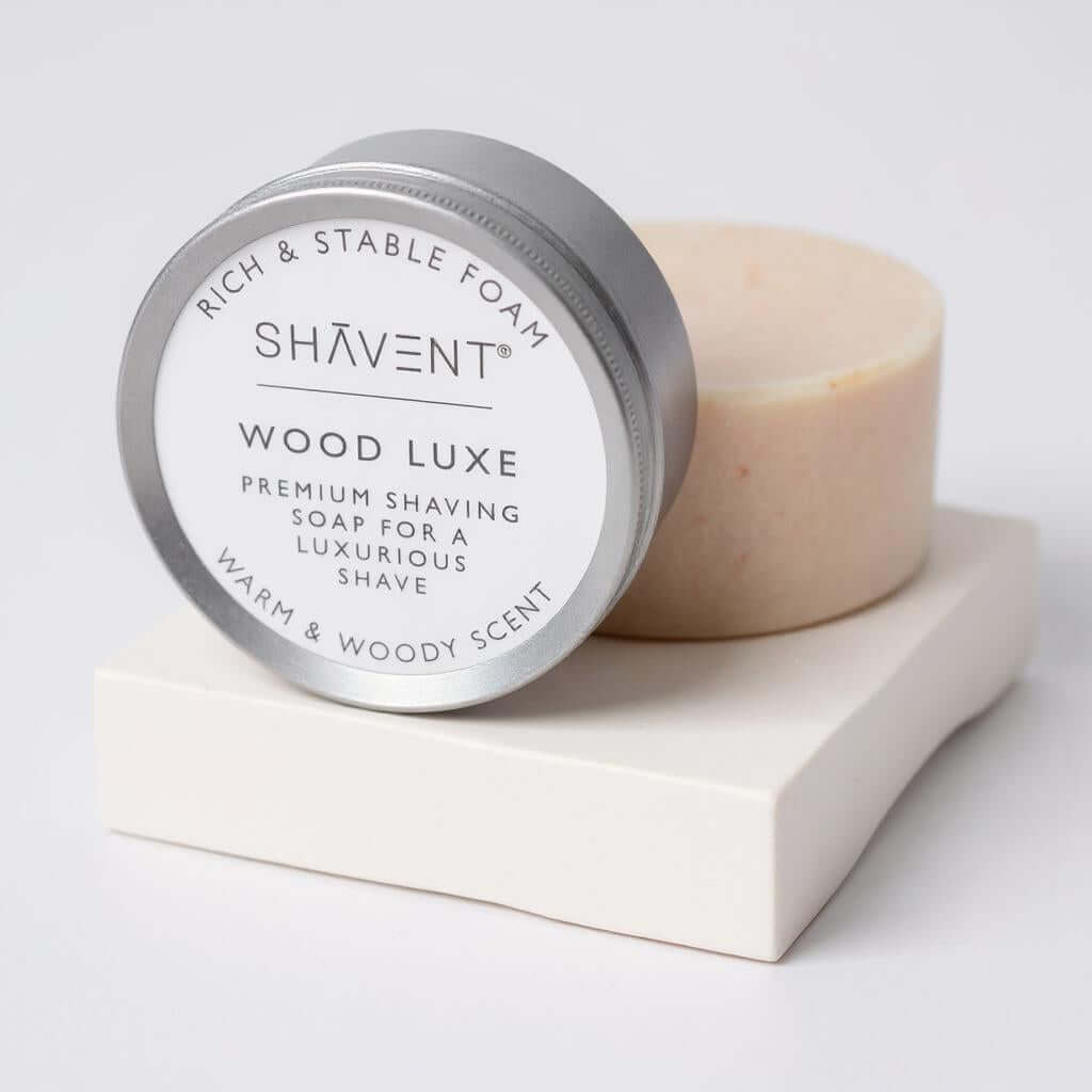 SHÄVENT Wood Luxe Premium Rasierseife im runden Blech, Rich & Stable Foam und Warm & Woody Scent, neben SeifenstĂŒck auf weiĂem Block. Teil des SHAVENT MĂ€nner-Geschenk-Sets.